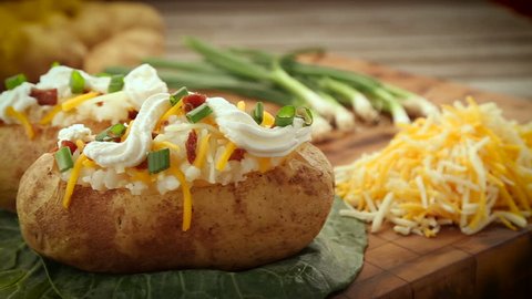 Baked potatoes fully garnished with chive, sour cream, bacon and shredded cheese. Potatoes are sitting on wood chopping block surrounded by pile of spuds, green onions, and a pile of shredded cheese. 