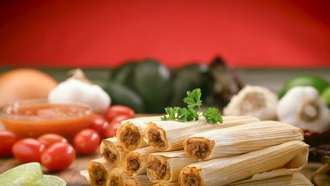 Tamales with husks stacked on a wood chopping board, surrounded by limes, cherry tomatoes, garlic cloves, salsa bowel, whole avocados and a yellow onion.