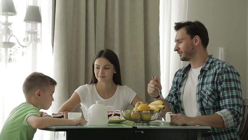Family of three having breakfast in the morning together in their home living room, smiling and with the mother pouring milk in a glass for the son, interior.