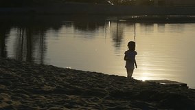 a Small Fair-Haired Girl Waving a Hand. She Looks Towards a Setting Sun. After That the Girl Sits Down and Starts to Consider a Shells Which Lie on the Ground. Video Fragment Was Recorded in