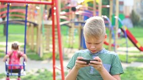 9 years old kid standing at public children playground playing computer games on cell phone. Anonymous parents and children in blurry background on summer day in busy city. Real time video footage