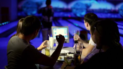 Young man makes strike in bowling game, his friends cheers hum and drink beer in dark club