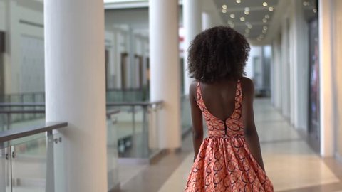 Young sexy afro-american girl walking in the bright orange dress in the trade centre, turning and looking back