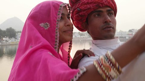 Pan left to a young Indian couple in traditional dresses taking a selfie with a phone mobile camera against the setting sun on Pushkar Lake, Rajasthan 