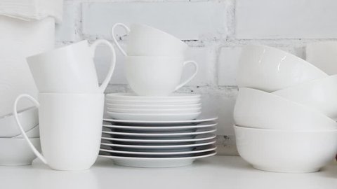 Stacked white porcelain dishes, plates, cups, saucers, mugs in restaurant kitchen interior. Slow pan. White background.