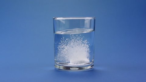 Aspirin or effervescent pill dropping into a glass of water on blue background.