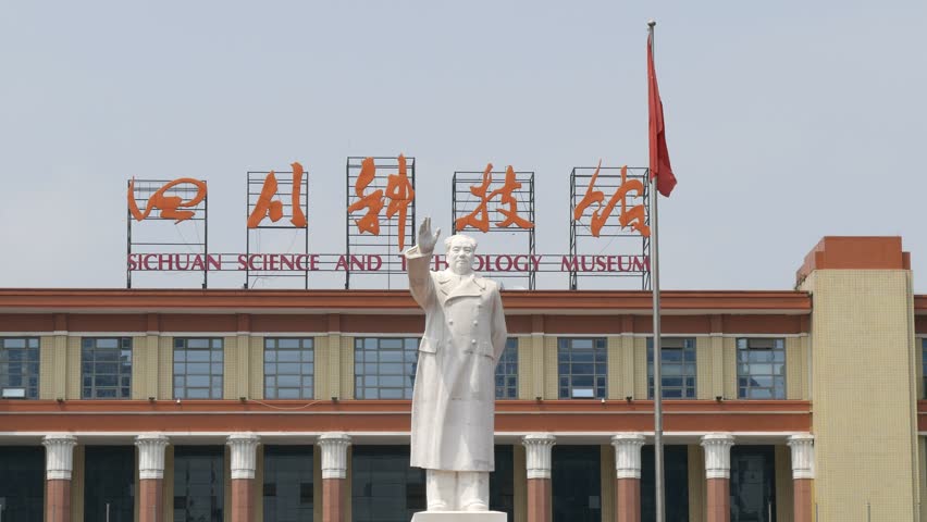 Close view of Chengdu Sichuan science & technology museum front roof line with English & Chinese sign, Chairman Mao statue and flag wrapped around flagpole with clear blue sky background.