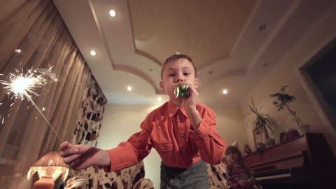 Little boy in Birthday cap holding sparkler and blowing whistle at camera in modern room.