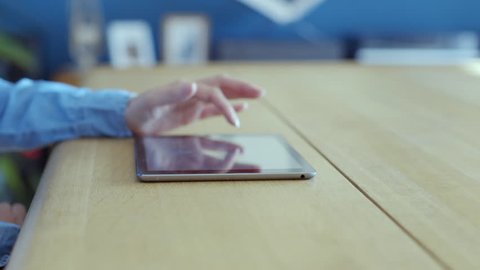 Panning shot of female hands using digital tablet. Midsection of woman in casual denim shirt is scrolling and zooming while online. She is sitting at the table in brightly lit house.