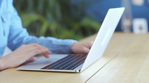 Panning shot of woman using touchpad on laptop. Female in casual denim shirt scrolling online while sitting at table in living room. She is in brightly lit home.