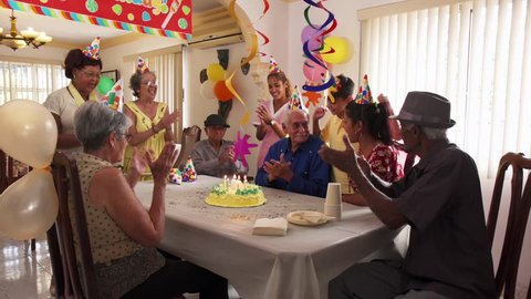 Group of old friends and family celebrating senior man birthday in retirement home. Happy elderly people having fun during party. Grandfather blowing candles on cake and smiling. Slow motion
