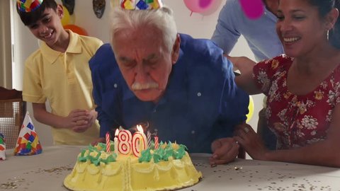 Group of old friends and family celebrating senior man birthday in retirement home. Happy elderly people having fun during party. Grandfather blowing candles on cake and smiling. Slow motion

