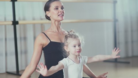 A woman and a girl in a ballet school. Beautiful and statuesque female ballerina doing stretches with a little girl. Adult ballerina practicing with the little girl and trains Russian classical ballet