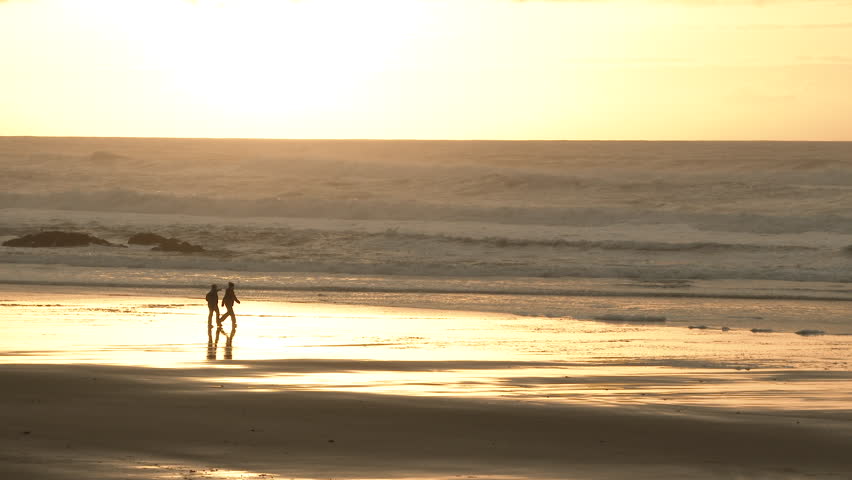 Two people walking along the Oregon Coast at sunset pass through frame while the Pacific produces rough seas.
