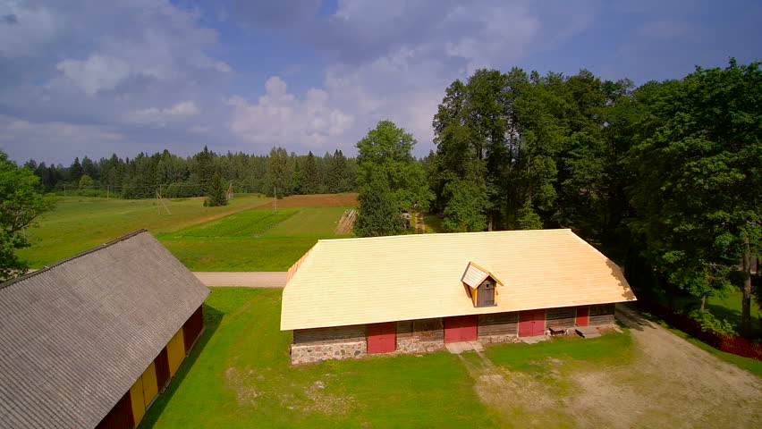 Aerial view of the cabin house in the field with the big trees on the back and the green lawn around