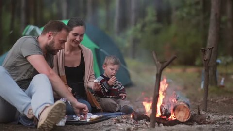 Family rest in the nature. Husband and wife with their little son are cooking marshmallows on open fire and eating them.