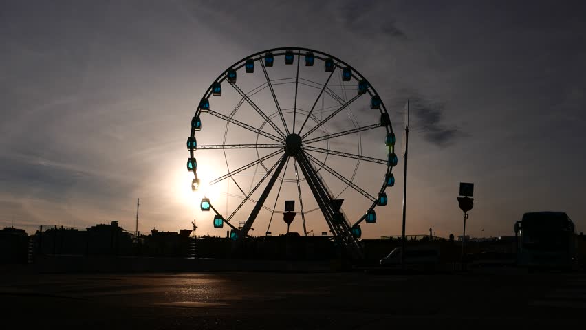 Silhouetted scene, man wheel on city bicycle against large Ferris Wheel, nice sunset scene high contrast shot. Bright sun spot shine in dusk sky. Bike rider approach and pass by, low angle camera