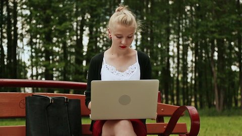 Beautiful young girl with blond hair on a park bench working on her laptop. Girl using laptop, typing. Front view.