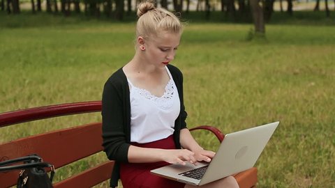 Beautiful young girl with blond hair on a park bench working on her laptop. Girl using laptop, typing. Side view.