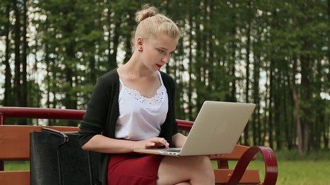 Beautiful young girl with blond hair on a park bench working on her laptop. Girl using laptop, typing.