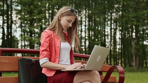 Beautiful young girl with blond hair on a park bench working on her laptop. Girl using laptop, typing.