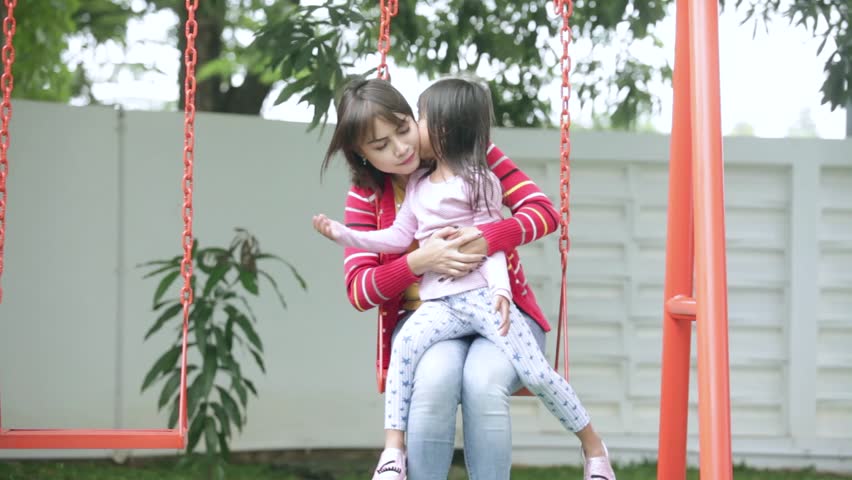 Young Asian mother sitting on a swing and kissing her daughter, shot at the playground