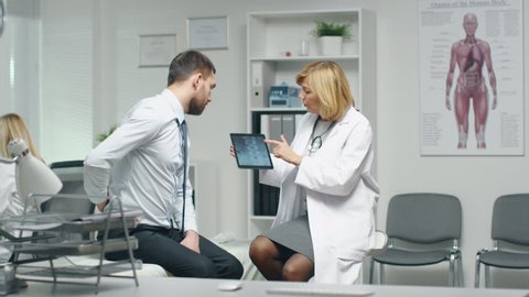 Mid Adult Female Doctor Consults Young Man About His Back Pain. Doctor Shows Him Tablet Computer With His Spine X-Ray.  