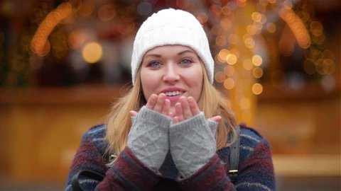 Happy Carefree Woman With Blonde Hair Sends An Air Kiss Over Christmas Lights Background. Beautiful Girl Wearing A White Hat And Grey Mitts Is Smiling On The New Year Fair.