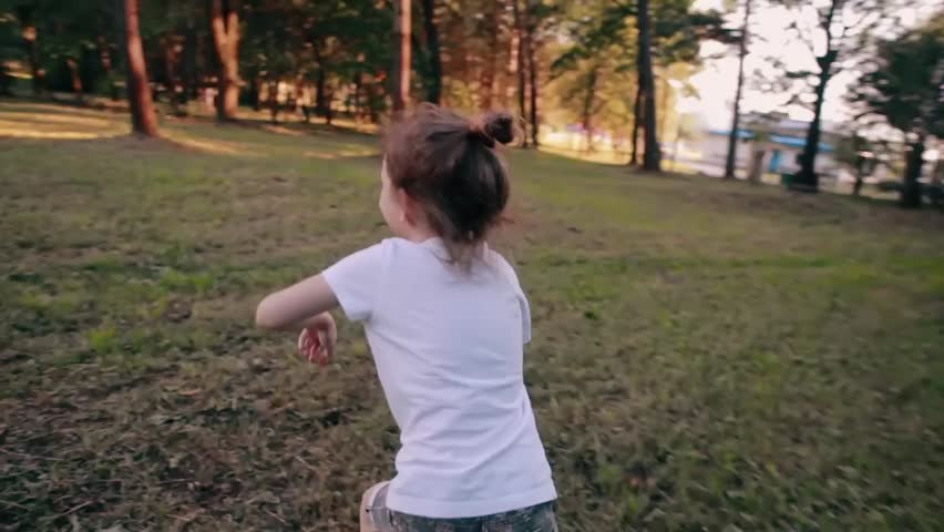 A cheerful little girl runs away from camera in a park on a sunny summer day. Back view, slow mo