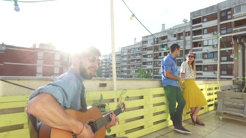 Musician playing guitar at rooftop party on sunny day, people hanging out in background