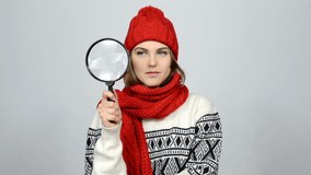 Searching winter woman. Girl wearing warm winter red knitted hat and scarf looking through magnifying glass at blank copy spaces at side and with amazement on the camera, studio shot