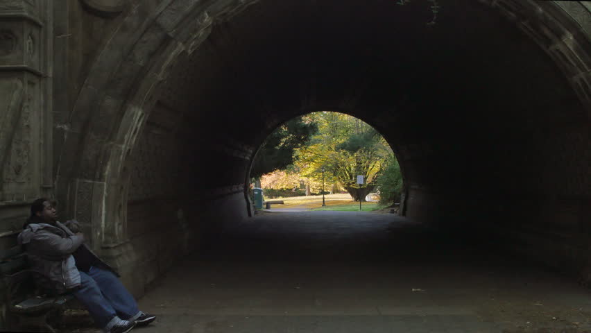 A Wide Shot of a Man Sitting in Front of a Bench at a Tunnel in a Park Using a Smart Phone