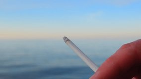Person smoking outdoor at the sea landscape background. Cigarette and smoke close-up, blue sky and sea on the soft blurred background. Full HD video footage