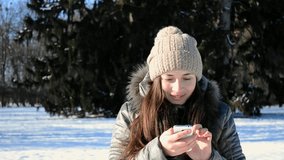 Young girl woman brunette woman with long hair typing a message on a smartphone winter day in the park. Close-up video. The girl looks into the camera and smiles broadly
