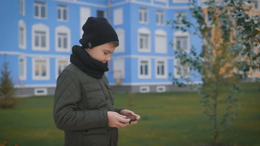 Child in warm dark grey jacket and black cap and scarf is playing phone standing in the street with buildings of different colours behind him. Young man is holding mobile phone in his hands