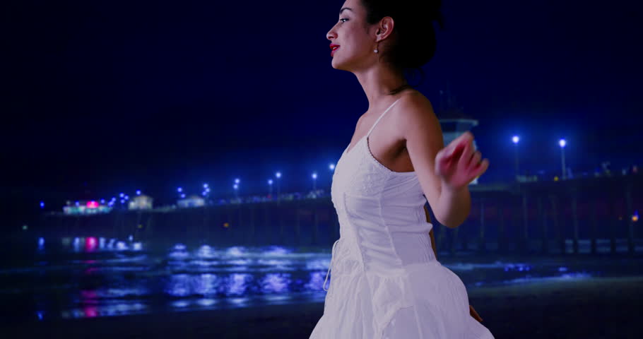 A Hispanic woman looks off on the waves, lit by a blue pier. A Latina girl stands near the waves lit by blue pier lights. 4k