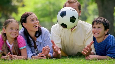 Smiling family trying to catch a soccer ball while lying in a park