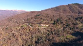 Aerial shot, little village in the middle of an autumn wood in the mountains with orange dry leaves, sun high in the sky, filmed with drone
