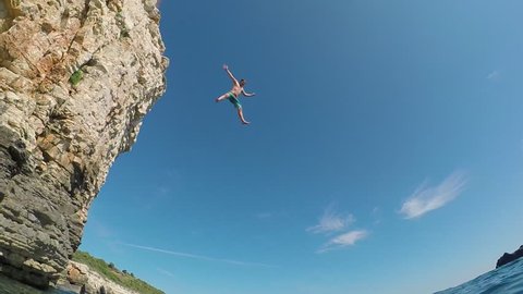 SLOW MOTION UNDERWATER: Happy young man with hands raised jumping into water off a rocky wall at sunny seaside. Fearless guy on fun summer vacation diving into refreshing sea off extremely high cliff