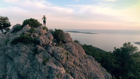 Aerial - Moving above young adult woman reaching the top of the seaside mountain at sunset
