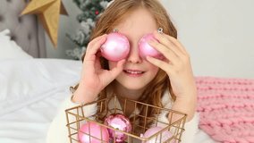Portrait of cute happy little girl on Christmas morning in home interior. Child paying with holiday Xmas decorations, making eye of pink balls while laying on bed. Real time full hd video footage.