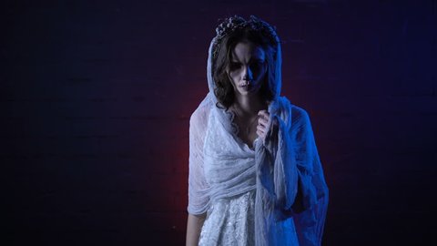 Ghost of lonely dead bride in white wedding dress and veil standing with sad face moving her hand. Scary girl halloween apperance make-up against horror background. Mysterious woman