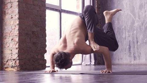 Young man doing an asana ashtavakrasana. Hand standing pose. Yogi master workout on black mat on urban studio. Guy doing yoga indoors near a window, lifestyle healthy concept