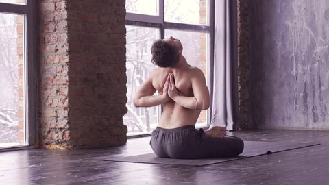 Young caucasian yoga master practicing yoga near a window. Yogi men sitting in lotus pose and meditates on dark urban background indoors. Yoga men workout in studio on black mat, back view