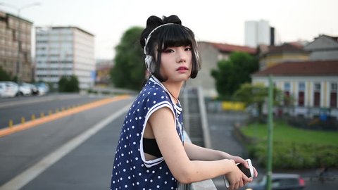 young beautiful asian millennial woman listening music with earphones and smart phone hand hold outdoor in city back light, looking at camera - relaxing, music, technology concept