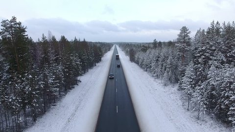 Camera back flying along winter road in evergreen woods of Karelia. Trucks driving on highway. The Kola route to the Murmansk, north of Russia
