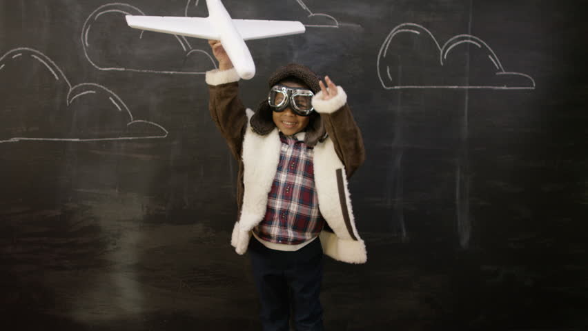 4K Cute little boy in pilot costume playing with toy plane in front of blackboard with drawings of clouds. Shot on RED Epic.