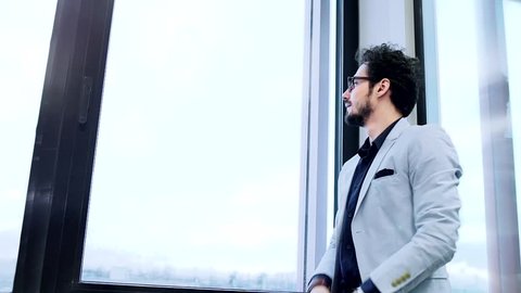 A young businessman stands at a large window in the office. Slow motion.