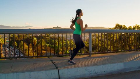 Athletic Woman Working Out. Jogging across a bridge. Trees, Sun and city can be seen in the background. Slow Motion.