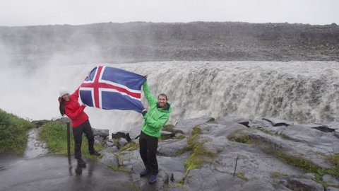 Travel couple fun by Dettifoss waterfall on Iceland showing Icelandic flag. People visiting famous tourist attractions and landmarks. Happy tourist couple enjoying vacation travel. 